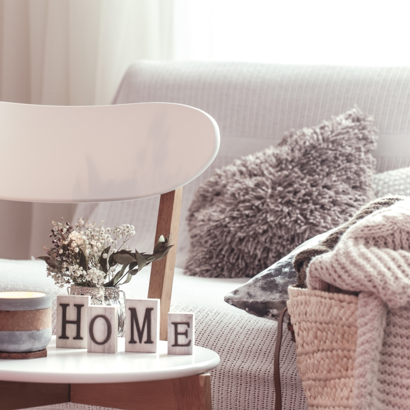Chic interior for a house. Candles, a vase with flowers with wooden letters of the home on wooden white chair. Sofa and wicker basket with cushions in the background. Home decoration.